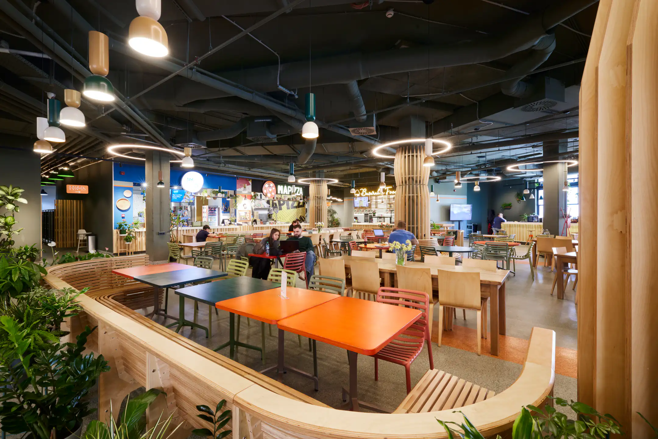 Food court area with curved wooden banquettes and orange square tables on a green platform