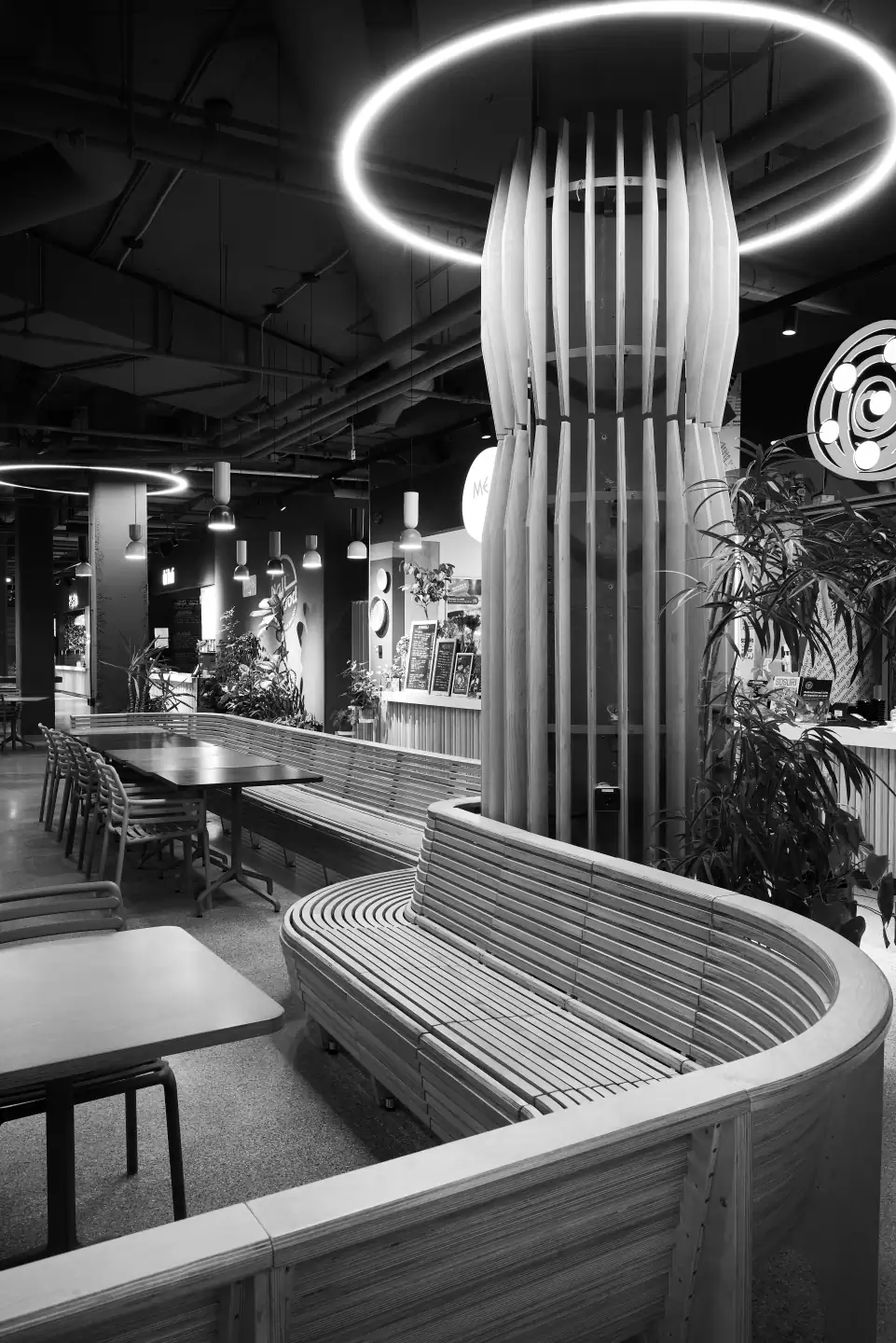 Sculptural wooden slat column lit by a circular ring fixture — black-and-white view of the food court